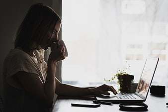 Woman drinking coffee while working on laptop by window