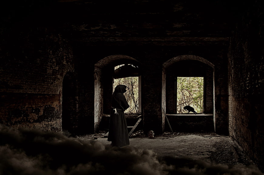 Dark hooded figure in abandoned ruin with fog and crow