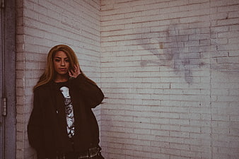 Young woman in black jacket leaning against white brick wall