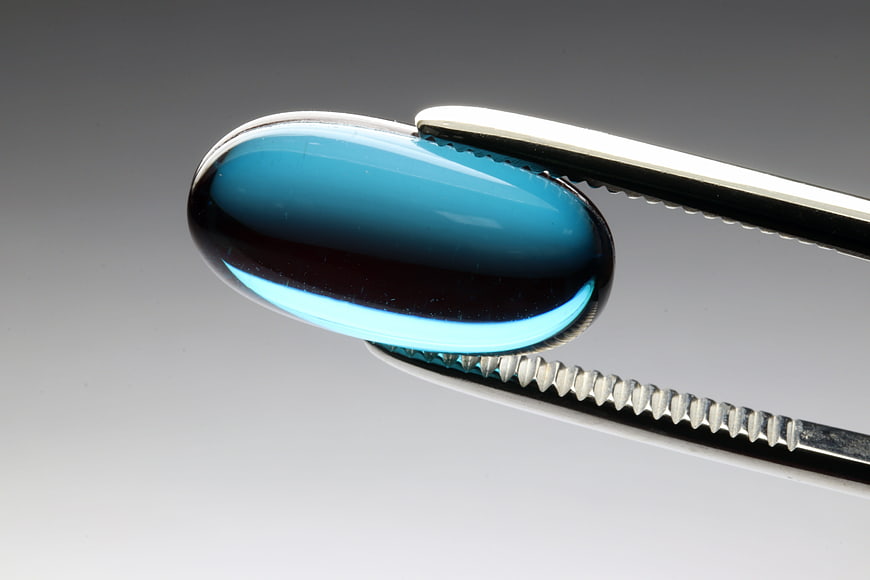 Blue Capsule Pill Held In Metal Tweezers On Gray Background