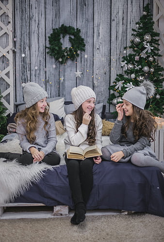 Three girls in winter hats reading book by Christmas tree