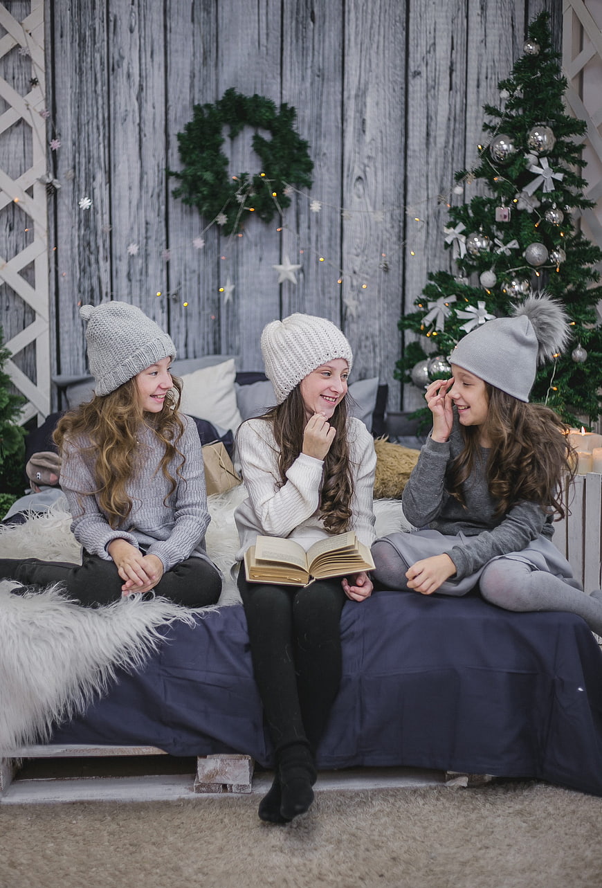 Three girls in winter hats reading book by Christmas tree