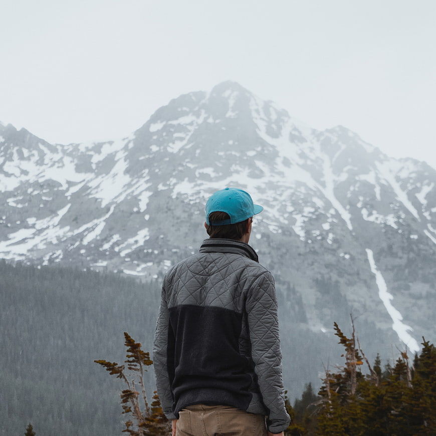 Man in Blue Cap Looking at Snowy Mountain Peak