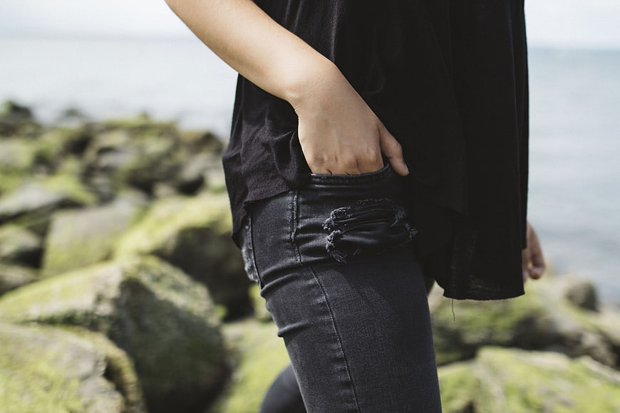 Woman in Black Jeans Standing on Mossy Rocks by Sea