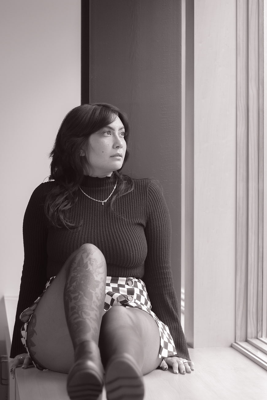 Black and white portrait of woman with tattoos sitting by window