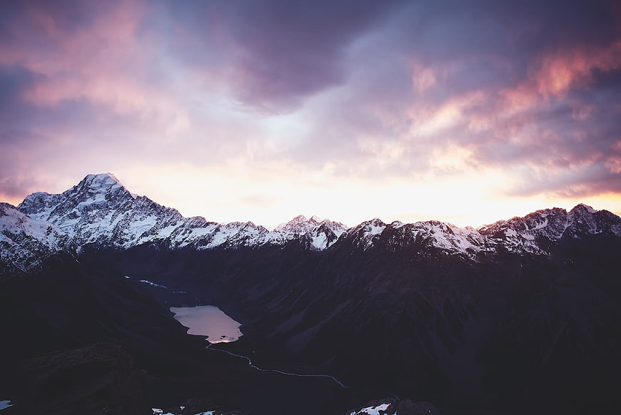 Snowy Mountain Range at Sunset with Purple Clouds and Lake