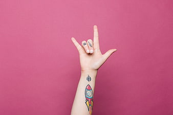 Hand with tattoos making rock sign on pink background