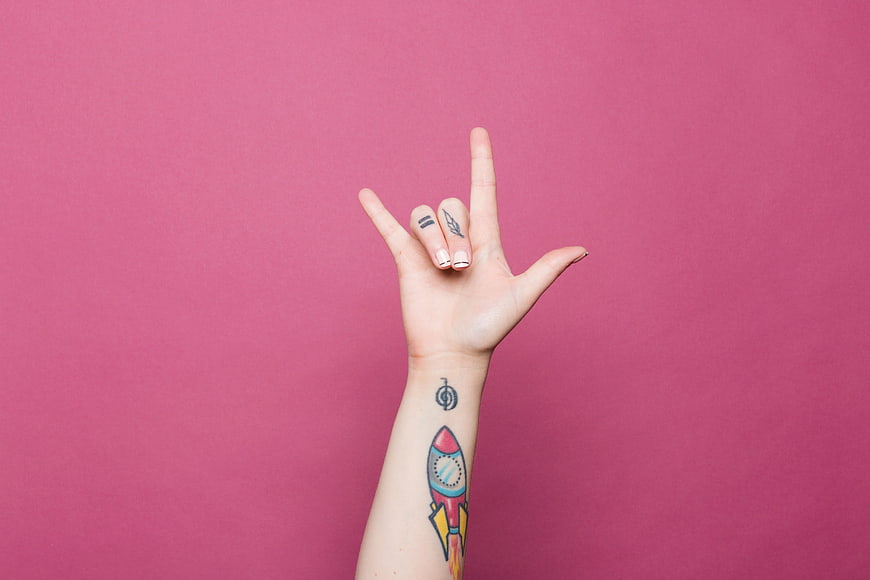 Hand with tattoos making rock sign on pink background