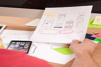 Hand holding paper wireframe sketches over smartphone on desk