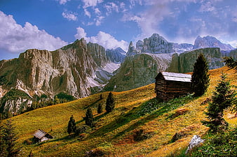 Rustic wooden cabin on grassy mountain slope with dramatic peaks