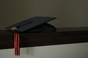 Black graduation cap with red tassel resting on a dark wooden shelf