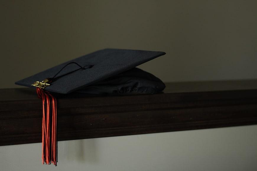 Black graduation cap with red tassel resting on a dark wooden shelf