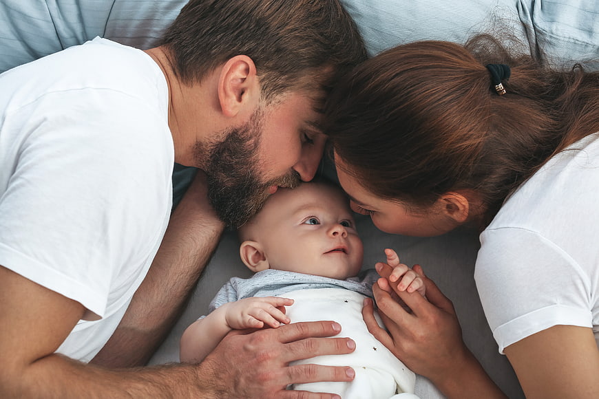Parents kissing baby on forehead in bed