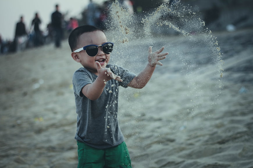 Happy toddler boy throwing sand at the beach wearing sunglasses