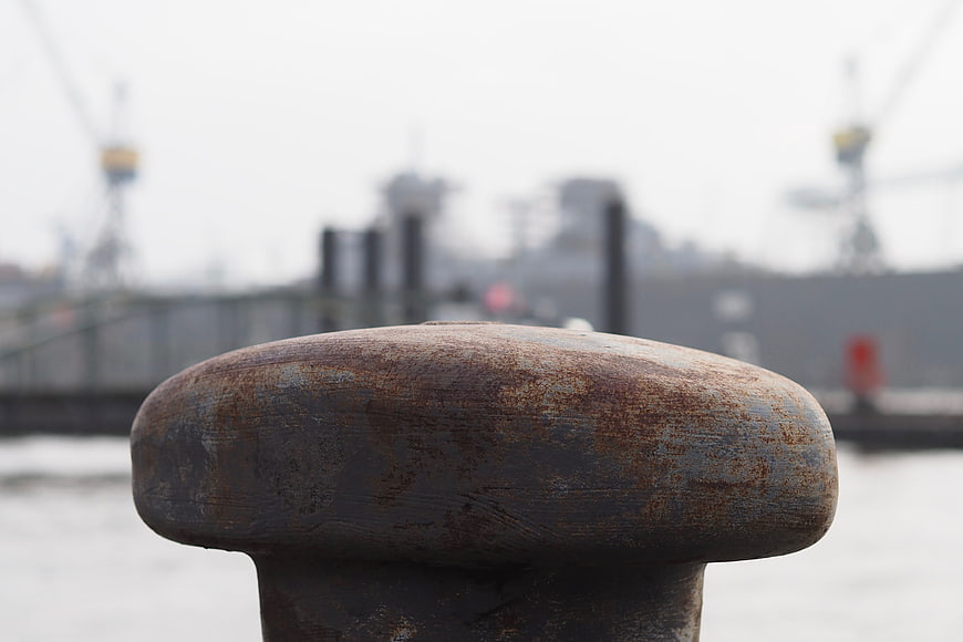 Close-up of rusty metal bollard at harbor with blurred ship background