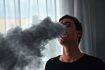 Young man exhaling thick vapor cloud indoors near window