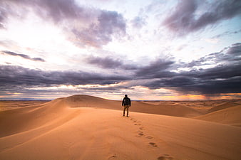 Lone Man Walking on Sand Dunes Under Dramatic Cloudy Sky