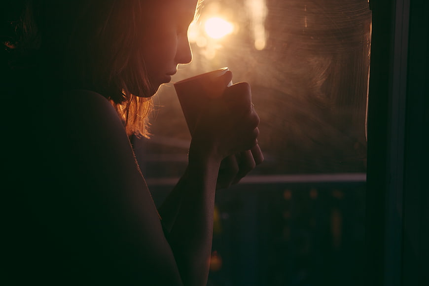 Woman drinking hot beverage by window at sunset