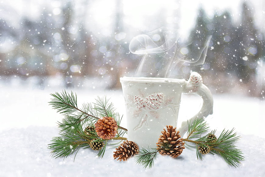 Steaming white mug with pine branches and cones in snow