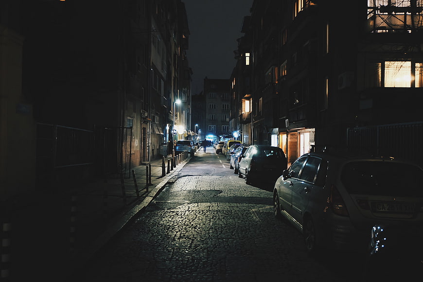 Dark cobblestone street at night with parked cars and city lights