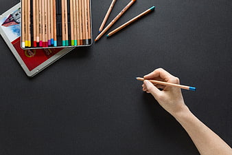 Hand holding colored pencil on black background with art supplies
