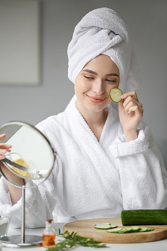 Woman in white bathrobe applying cucumber slice to face