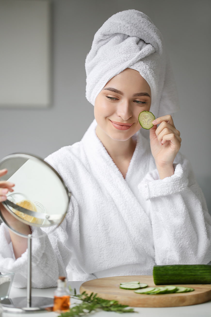 Woman in white bathrobe applying cucumber slice to face