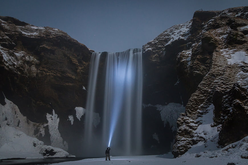 Man with Flashlight Under Snowy Waterfall at Night