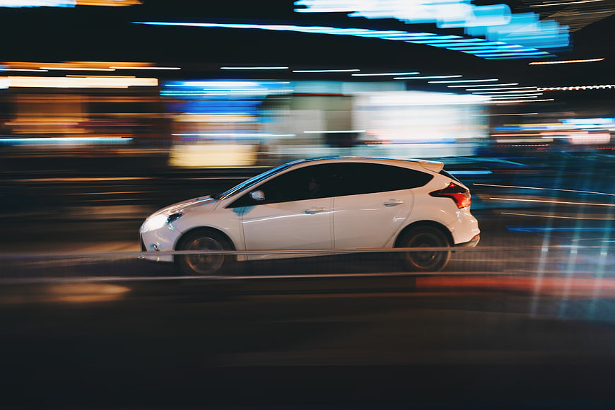 White car driving fast at night with city light trails