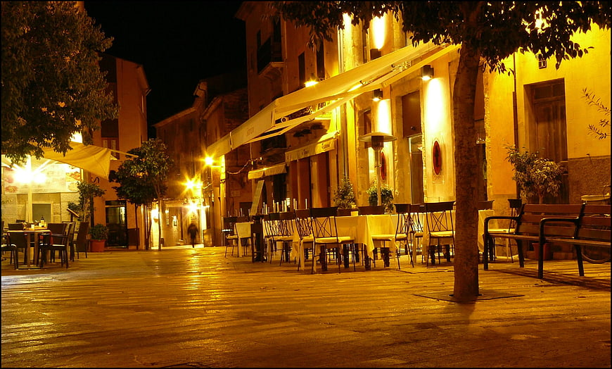 Illuminated European Village Square With Outdoor Bistro Seating At Night