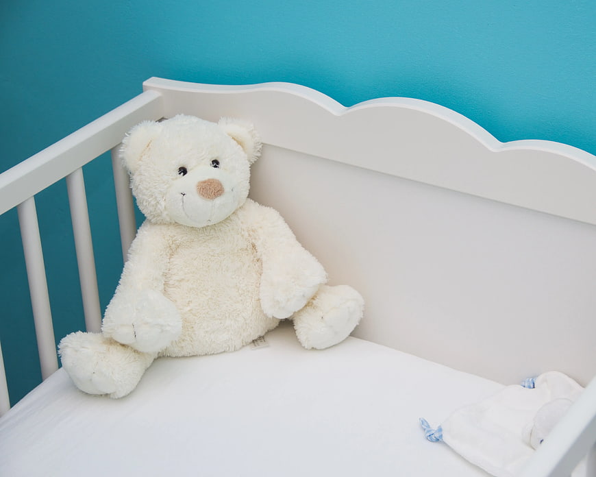 White teddy bear sitting in a white crib against blue wall