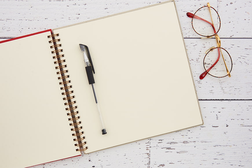 Flat lay of blank notebook, pen, and glasses on white desk