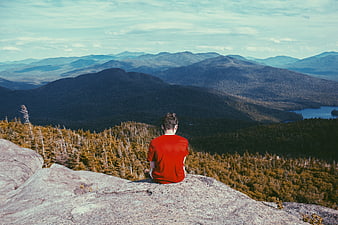 Man in Red Shirt Sitting on Mountain Summit Overlooking Forest