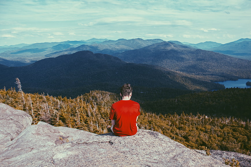 Man in Red Shirt Sitting on Mountain Summit Overlooking Forest