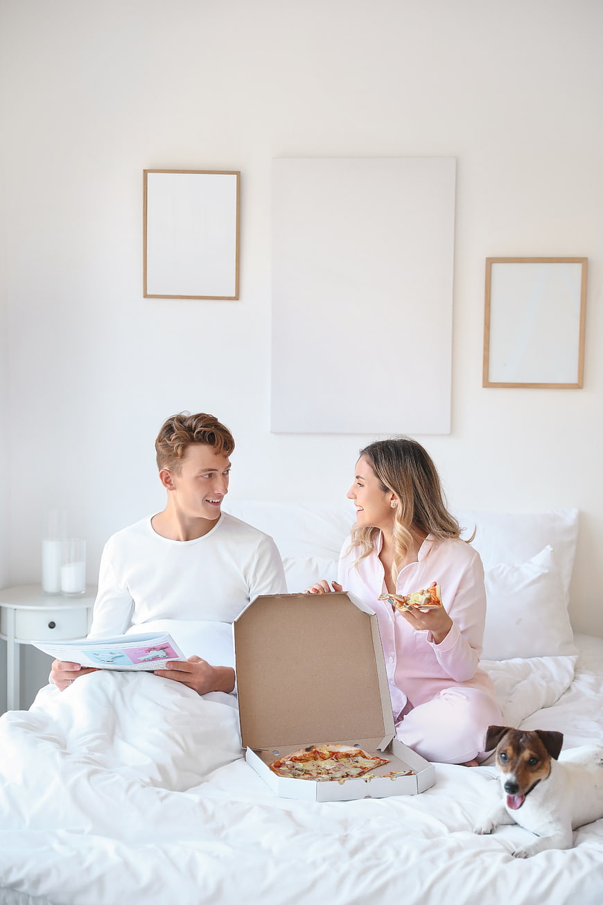 Happy couple eating pizza in bed with dog