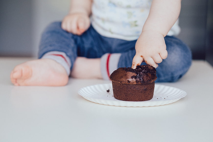 Baby Reaching For Chocolate Muffin On White Plate