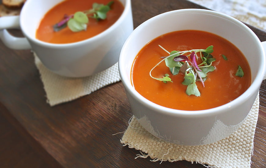 Two bowls of creamy tomato soup garnished with fresh microgreens on a wooden table