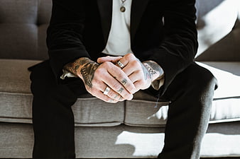 Tattooed man in black suit sitting on sofa with clasped hands