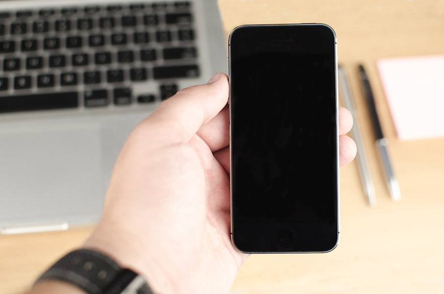 Hand holding black smartphone with blank screen over laptop workspace