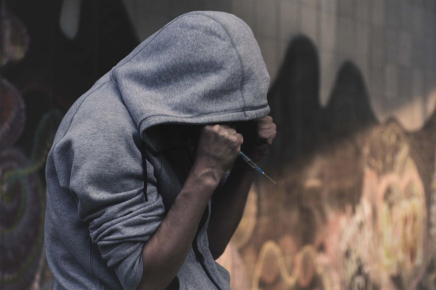 Hooded figure holding syringe against graffiti wall