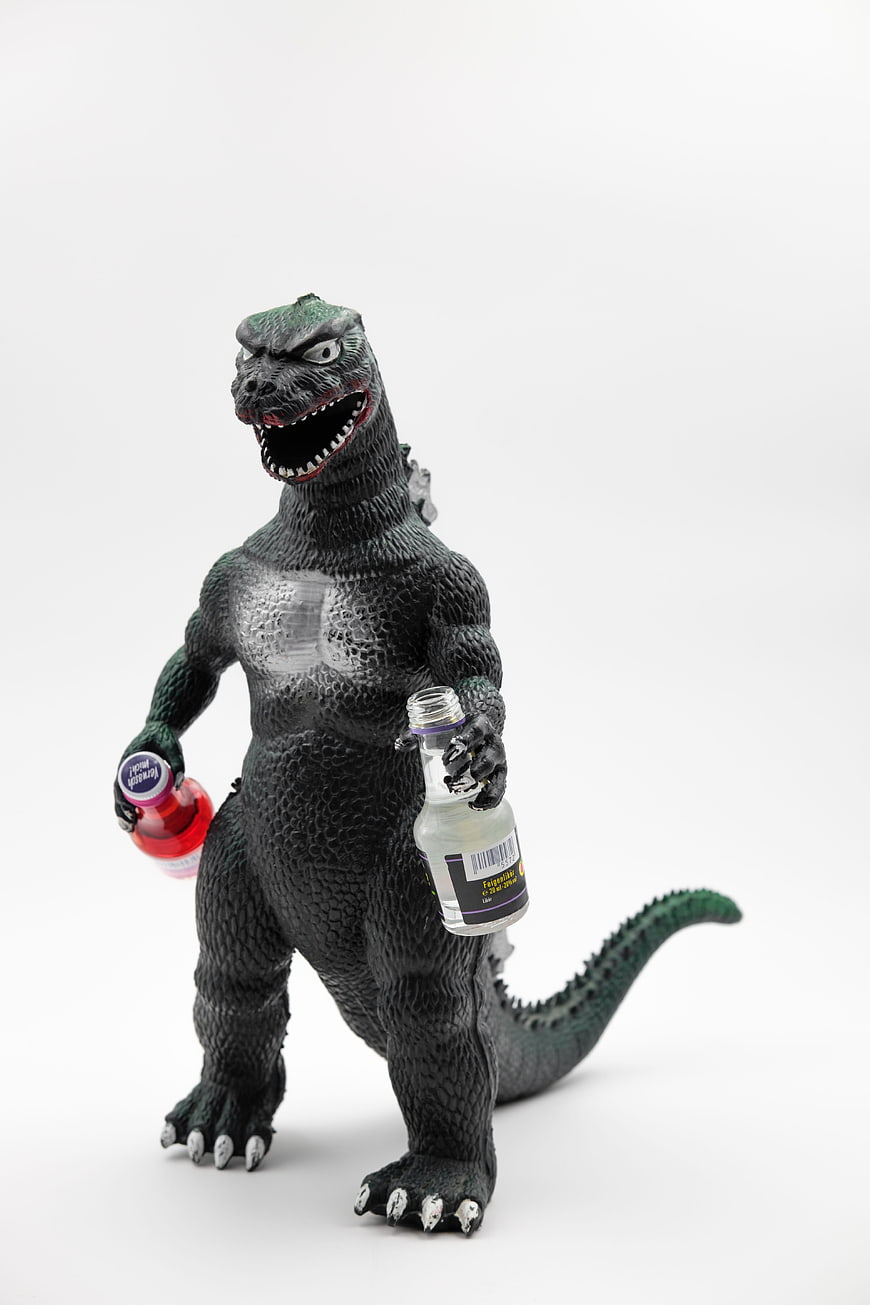 Godzilla toy figure holding two small liquor bottles on white background