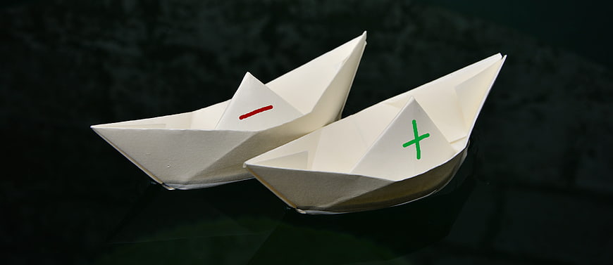 Two paper boats with plus and minus signs on dark water
