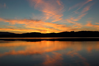 Serene Lake Sunset With Vibrant Orange Clouds And Mountain Silhouettes