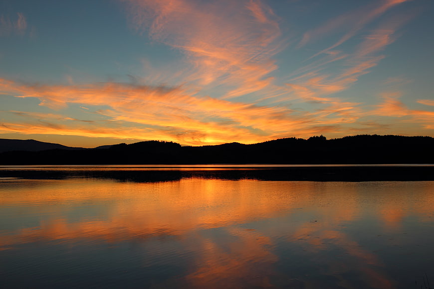 Serene Lake Sunset With Vibrant Orange Clouds And Mountain Silhouettes