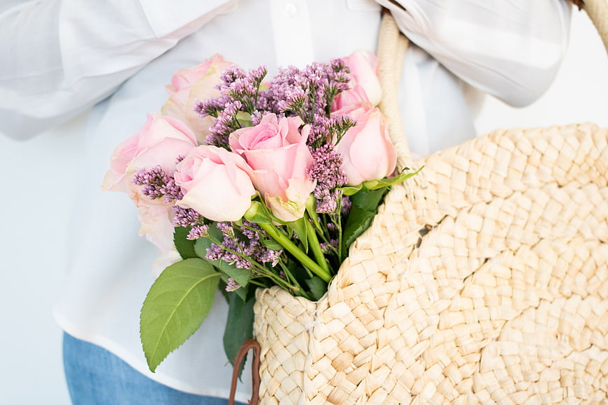 Woman holding pink roses and purple flowers in straw tote