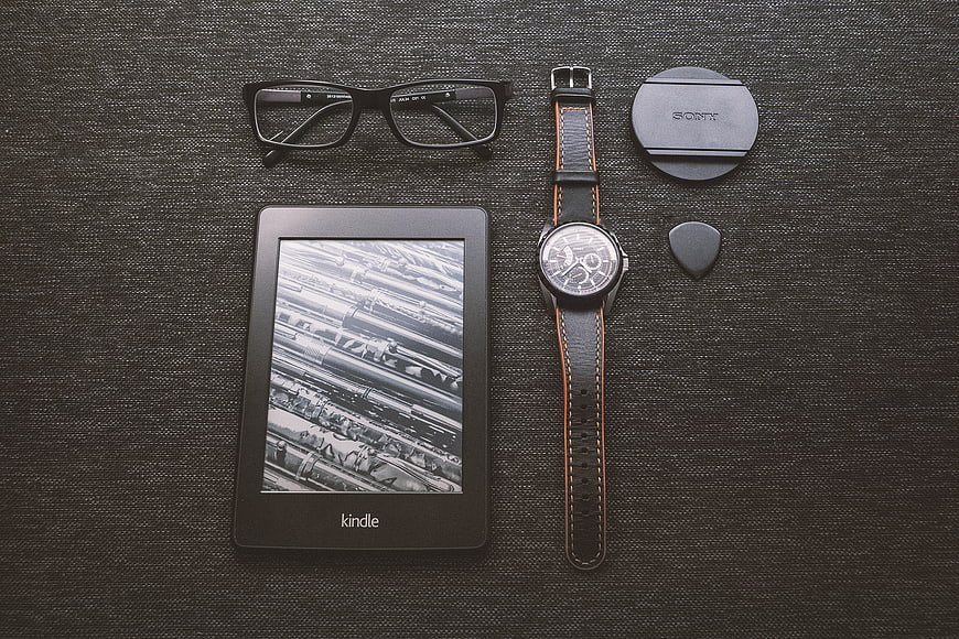 Flat lay of Kindle e-reader, watch, glasses, and camera lens cap