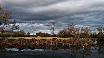Dramatic cloudy sky over riverbank with young trees and reflections