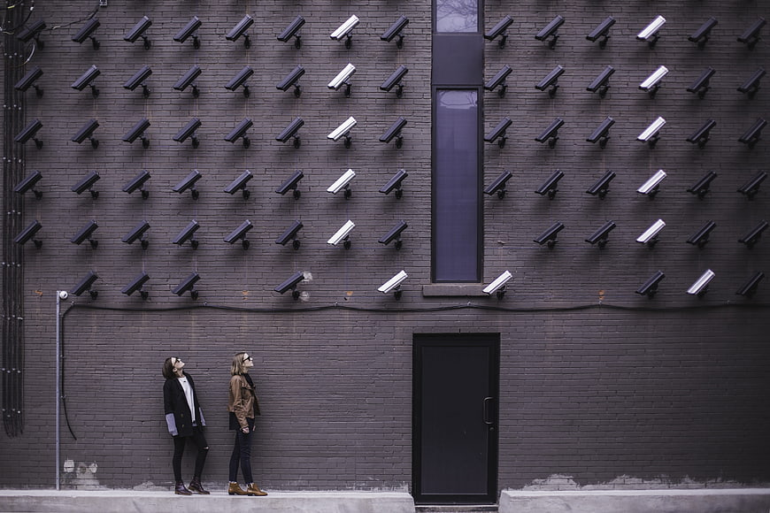 Two women standing under rows of security cameras on gray wall