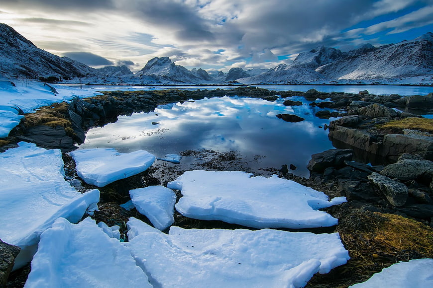 Snowy ice chunks on rocky shore with mountain reflection