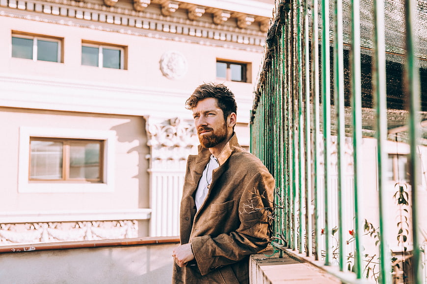 Handsome Bearded Man in Brown Coat Leaning on Green Railing